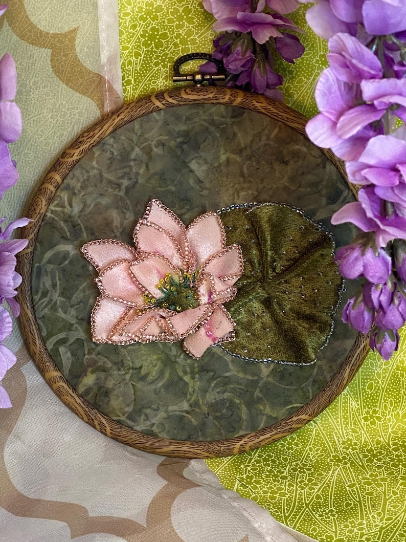 May include: A handcrafted embroidery hoop featuring a pink lotus flower with gold accents and a green velvet leaf. The artwork is set against a mottled green background within a wooden hoop, with purple flowers in the background.