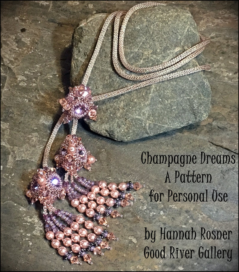 K&ouml;nnte beinhalten: Eine Perlenkette mit zwei rosa und violetten Quastenanh&auml;ngern. Die Kette besteht aus einer silbernen Kette und die Quasten sind aus Perlen und Kristallen gefertigt. Die Kette liegt auf einem grauen Stein mit dem Text "Champagne Dreams A Pattern for Personal Use by Hannah Rosner Good River Gallery".