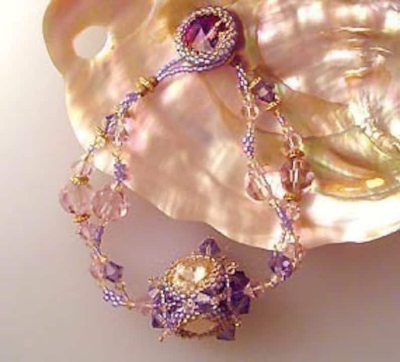 K&ouml;nnte beinhalten: Ein Armband mit Perlen und einem violetten und wei&szlig;en Kristall als Mittelpunkt. Das Armband ist aus klaren und rosafarbenen Perlen gefertigt und hat einen goldenen Akzent.
