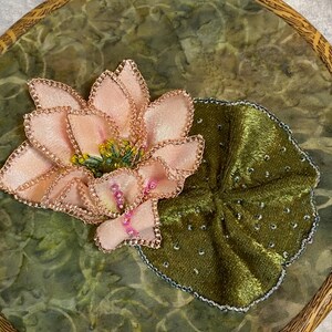 May include: Embroidered artwork featuring a pink lotus flower and a green lily pad. The flower has delicate petals with gold trim and a yellow center. The lily pad is a deep green with silver accents, set on a green background.