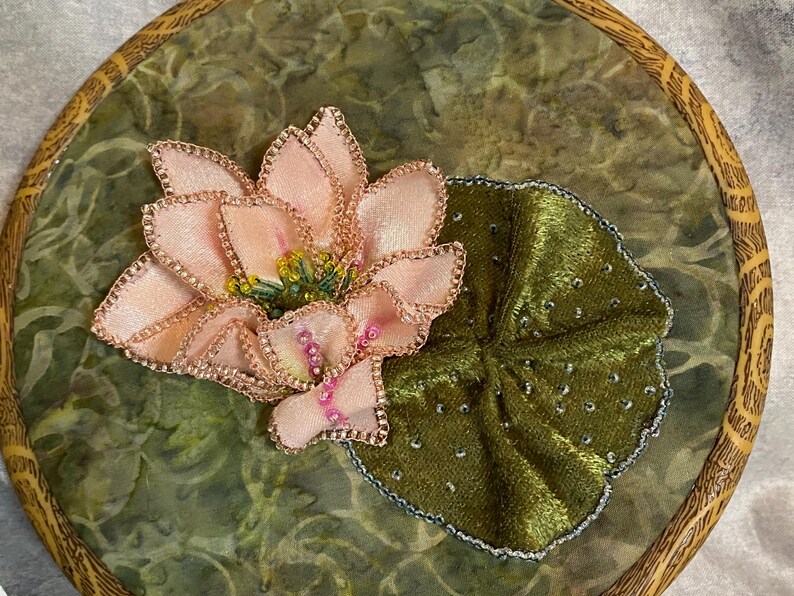 May include: Embroidered artwork featuring a pink lotus flower and a green lily pad. The flower has delicate petals with gold trim and a yellow center. The lily pad is a deep green with silver accents, set on a green background.