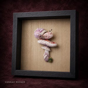 May include: A framed textile art piece featuring a three-dimensional embroidered snail resting on a mushroom. The snail has a pink and white shell, while the mushroom has a red and white cap with a textured stem. The text HANNAH ROSNER is visible.