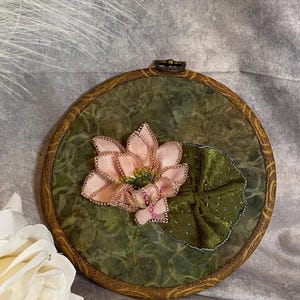 May include: A handcrafted embroidery piece featuring a pink lotus flower and a green lily pad. The flower has delicate pink petals with gold accents, and the lily pad is a rich green. The artwork is set in a round wooden hoop.