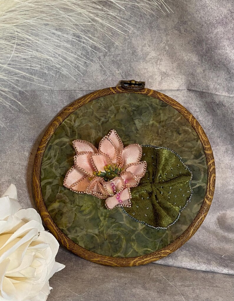 May include: A handcrafted embroidery piece featuring a pink lotus flower and a green lily pad. The flower has delicate pink petals with gold accents, and the lily pad is a rich green. The artwork is set in a round wooden hoop.