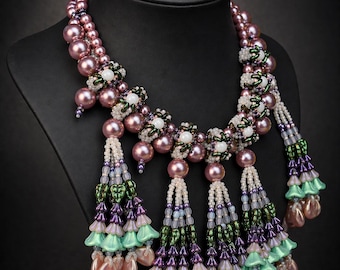 May include: A handcrafted necklace with a layered design, featuring pink pearl beads and beaded floral accents. The necklace has multiple beaded tassels in shades of purple, green, and pink. The necklace is displayed on a black mannequin.