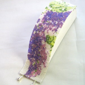 Op de afbeelding: Een kralenarmband met een bloemenpatroon in tinten paars, groen en wit. De armband is gemaakt met kleine kraaltjes en heeft zilveren sluitingen.