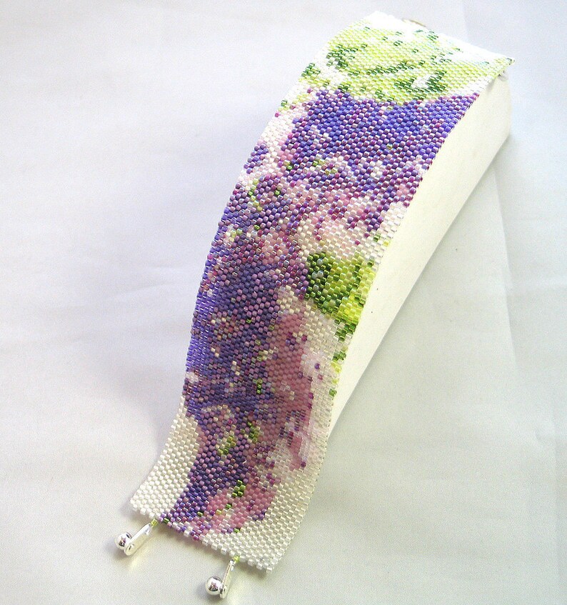 Op de afbeelding: Een kralenarmband met een bloemenpatroon in tinten paars, groen en wit. De armband is gemaakt met kleine kraaltjes en heeft zilveren sluitingen.