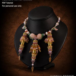 May include: A beaded necklace titled Make Love Not War Necklace PDF Tutorial For personal use only. The piece features three gold-toned cylindrical components adorned with intricate pink, yellow, and gold seed beads, accented by dangling leaf-shaped charms and pale pink stone elements.
