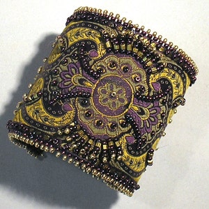 May include: A wide purple and gold fabric cuff bracelet with a paisley pattern and beaded trim.