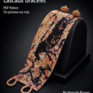 May include: A wide beaded bracelet titled Lascaux Bracelet by Hannah Rosner, featuring a complex pattern of black, orange, pink, and cream seed beads. The design includes two small beaded spheres at the top and two beaded loops at the bottom.