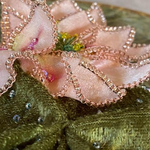 May include: Close-up of a decorative floral embellishment. The flower has soft pink petals with beaded edges and a yellow center. It sits atop a textured, olive green fabric, adorned with scattered iridescent beads.