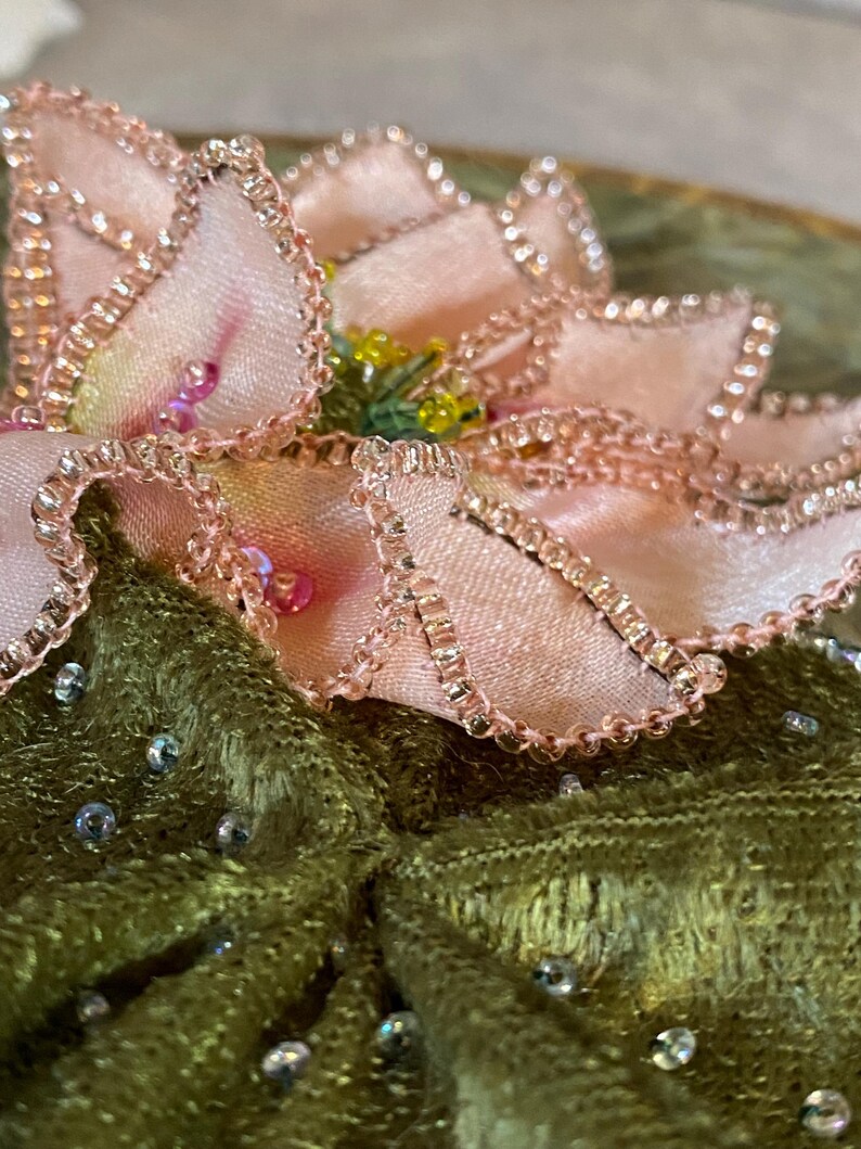 May include: Close-up of a decorative floral embellishment. The flower has soft pink petals with beaded edges and a yellow center. It sits atop a textured, olive green fabric, adorned with scattered iridescent beads.