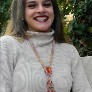 May include: A beaded necklace with a large orange gemstone pendant and a tassel of beads. The necklace is made of pink and gold beads and is worn over a light beige turtleneck sweater. The necklace is a statement piece.