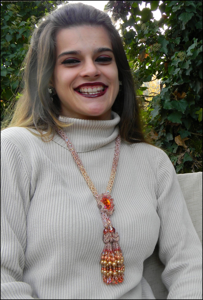 May include: A beaded necklace with a large orange gemstone pendant and a tassel of beads. The necklace is made of pink and gold beads and is worn over a light beige turtleneck sweater. The necklace is a statement piece.