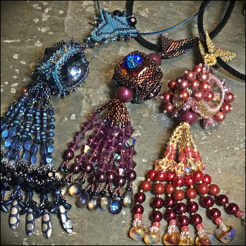 May include: Three beaded necklaces with crystal and pearl accents. Each necklace features a unique pendant with a tassel. The pendants are a blue geometric shape, a purple geometric shape, and a red geometric shape.