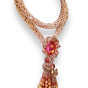 May include: A handcrafted necklace featuring a woven, rose-gold colored chain. The pendant showcases a large, faceted pink gemstone surrounded by smaller gems. A beaded tassel of gold and pink beads hangs below.