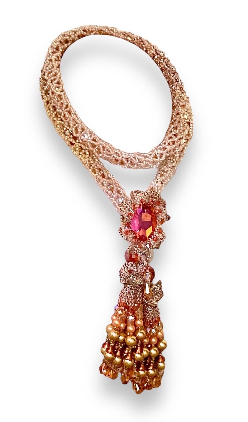 May include: A handcrafted necklace featuring a woven, rose-gold colored chain. The pendant showcases a large, faceted pink gemstone surrounded by smaller gems. A beaded tassel of gold and pink beads hangs below.