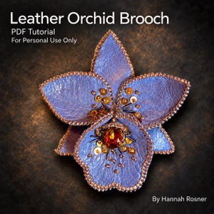Può includere: Un tutorial in PDF per una spilla a forma di orchidea in pelle di Hannah Rosner. La spilla presenta petali testurizzati color viola chiaro bordati con perline metalliche, centrati con un grande cristallo ambrato, paillettes dorate e accenti metallici scuri.