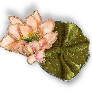 May include: A handcrafted, fabric lotus flower with a green leaf. The flower has soft pink petals with gold trim and a yellow center. The leaf is a deep green with silver accents. This floral accessory is perfect for adding a touch of nature to any outfit.