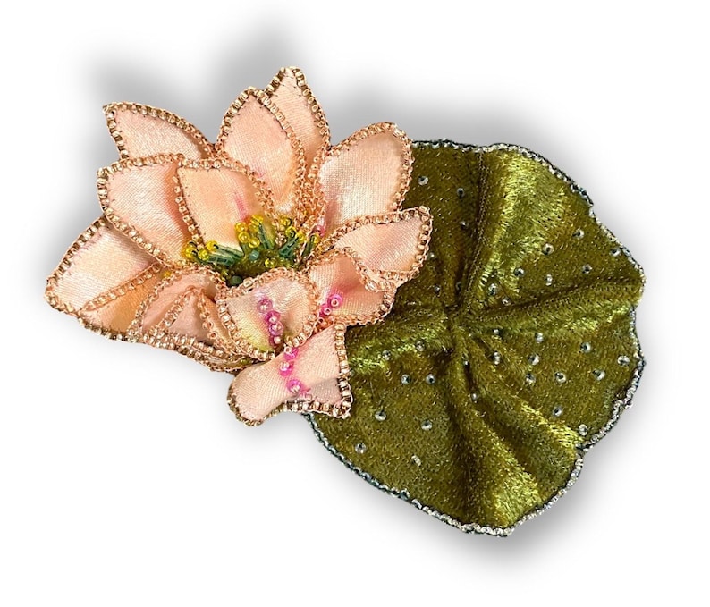 May include: A handcrafted, fabric lotus flower with a green leaf. The flower has soft pink petals with gold trim and a yellow center. The leaf is a deep green with silver accents. This floral accessory is perfect for adding a touch of nature to any outfit.