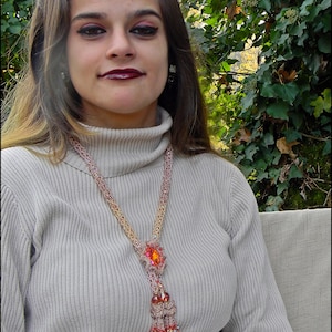 May include: A beaded necklace with a large orange gemstone pendant and a tassel of beads. The necklace is made of pink, gold, and orange beads. The person is wearing a light gray turtleneck sweater.