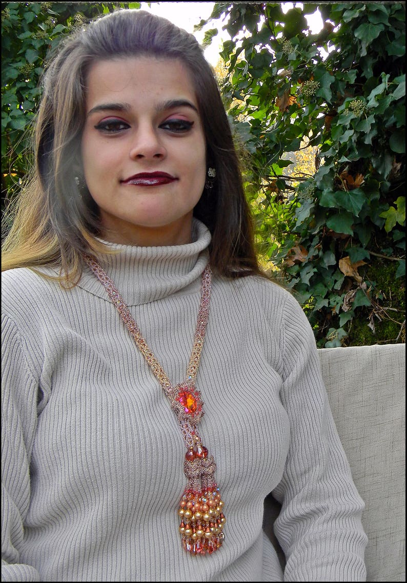 May include: A beaded necklace with a large orange gemstone pendant and a tassel of beads. The necklace is made of pink, gold, and orange beads. The person is wearing a light gray turtleneck sweater.