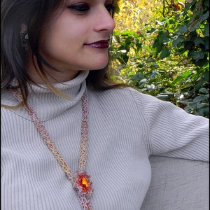 May include: A beaded necklace with a large orange gemstone pendant and a tassel. The necklace is made of small, colorful beads in shades of pink, gold, and orange. The woman is wearing a light gray turtleneck sweater.