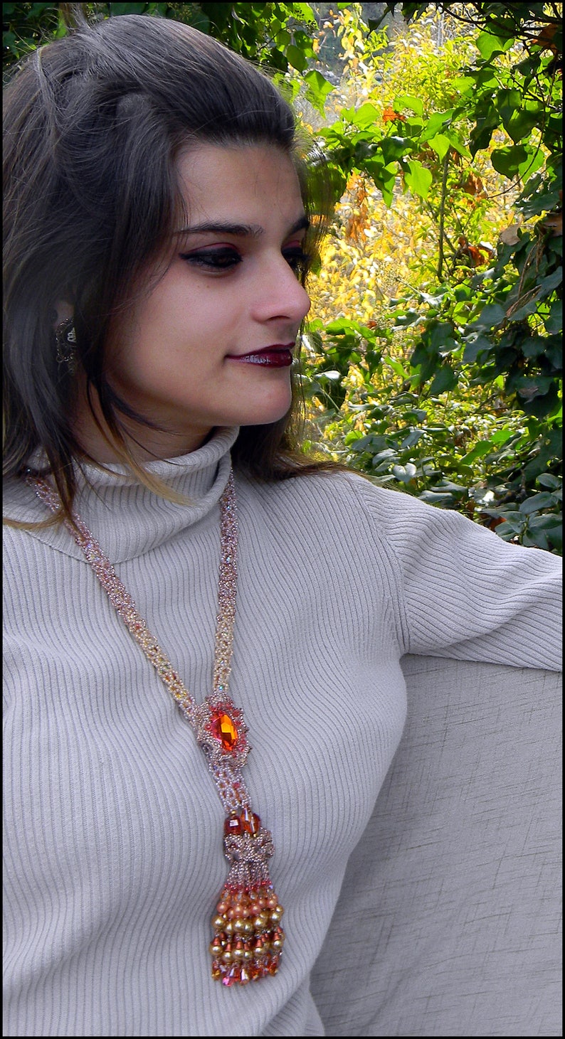 May include: A beaded necklace with a large orange gemstone pendant and a tassel. The necklace is made of small, colorful beads in shades of pink, gold, and orange. The woman is wearing a light gray turtleneck sweater.