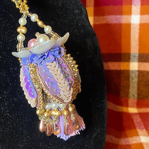May include: A handcrafted pendant necklace featuring an owl design. The owl is decorated with gold beads, pink and purple sequins, and pearl accents. A pink ribbon and gold chain complete the necklace. The necklace is a unique accessory.