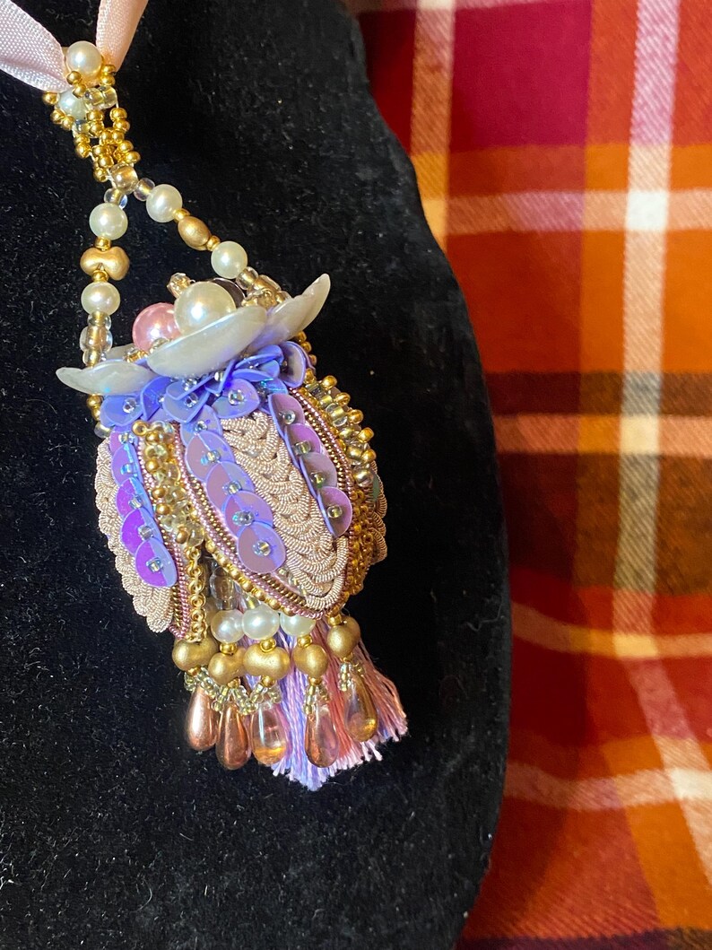 May include: A handcrafted pendant necklace featuring an owl design. The owl is decorated with gold beads, pink and purple sequins, and pearl accents. A pink ribbon and gold chain complete the necklace. The necklace is a unique accessory.