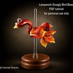 Op de afbeelding: Een lampwork glazen vogelkraal met wervelende rode, oranje en gele patronen, voorzien van wiebeloogjes en een gele snavel. De vogel is gemonteerd op een doorzichtige glazen staaf op een ronde houten voet. Tekst: Lampwork Googly Bird Bead PDF tutorial for personal use only. By Hannah Rosner.
