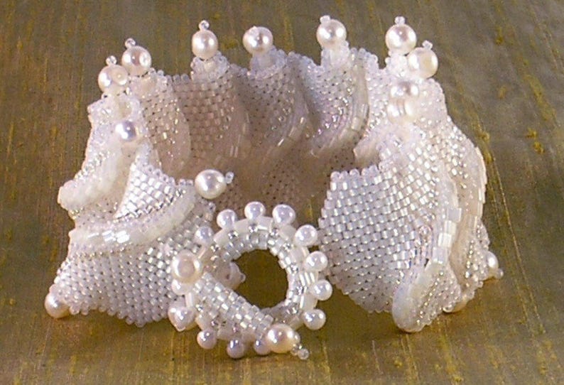 Puede incluir: Un brazalete de cuentas blancas con un dise&ntilde;o delicado e intrincado. El brazalete est&aacute; hecho de peque&ntilde;as cuentas blancas que se tejen juntas para crear una forma fluida y org&aacute;nica. El brazalete est&aacute; acentuado con cuentas blancas m&aacute;s grandes que a&ntilde;aden un toque de brillo.
