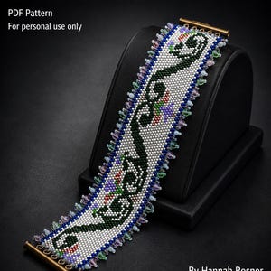 Op de afbeelding: Een brede kralenarmband met een witte achtergrond en een donkergroen vloeiend patroon, geaccentueerd door kleine blauwe, paarse en rode kralen. De randen zijn afgewerkt met iriserende kralen en goudkleurige metalen staven. Tekst: Art Nouveau Bracelet.