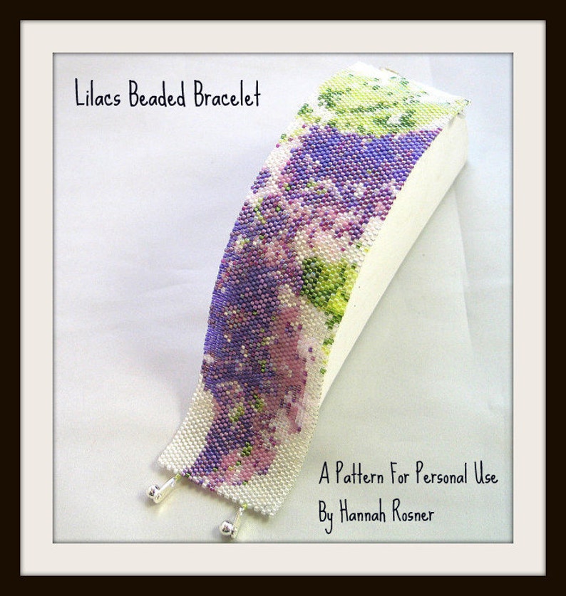 Op de afbeelding: Lila Perlen Armband, een kralenarmband met een paars, groen en wit bloemenpatroon. De armband ligt op een wit oppervlak. Een patroon voor persoonlijk gebruik door Hannah Rosner.