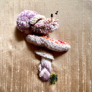 May include: A pink and white beaded snail with black eyes sits on top of a mushroom with a brown cap and a white stem. The mushroom is on top of a larger mushroom with a red and white cap and a white stem.