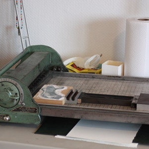 May include: A green metal paper trimmer with a dial and a lever. The trimmer is on a wooden table with a white paper pad and a black mat.