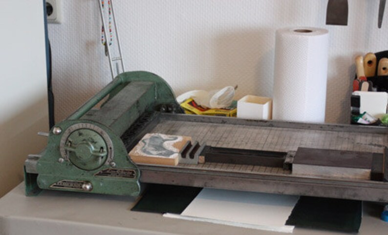 May include: A green metal paper trimmer with a dial and a lever. The trimmer is on a wooden table with a white paper pad and a black mat.