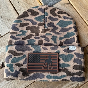 May include: A brown and green camouflage beanie with a brown leather patch featuring a black silhouette of a dog and an American flag. The patch also has the text "LOST" on it. The beanie has the text "THE BLANKS YOU'VE ALWAYS WANTED" on a tag attached to it.