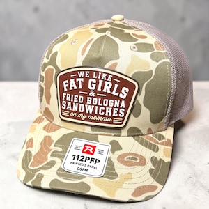 May include: A camouflage trucker hat with a brown patch featuring the white text WE LIKE FAT GIRLS & FRIED BOLOGNA SANDWICHES on my momma. The hat has a mesh back and a sticker on the brim that reads EST 1970 R 112PFP PRINTED 5 PANEL OSFM.