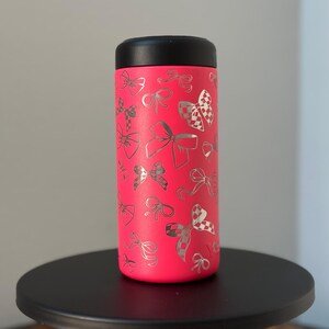 以下が含まれることがあります： 黒い蓋が付いたピンクのステンレス製タンブラー。タンブラーには、全体に銀色のリボン模様が施されています。