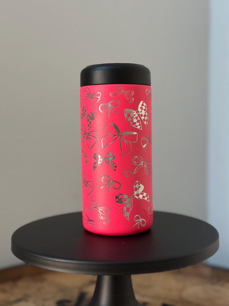 以下が含まれることがあります： 黒い蓋が付いたピンクのステンレス製タンブラー。タンブラーには、全体に銀色のリボン模様が施されています。