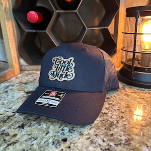 May include: Navy blue trucker hat with the text "Cool Little Dude" in a cursive font. The hat has a mesh back and a curved brim. The Richardson brand tag is visible.