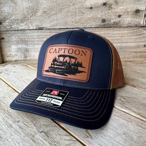 May include: Navy blue and brown trucker hat with a leather patch. The patch features the word "CAPTOON" above a pontoon boat graphic. The hat has a curved brim and a mesh back. The brand "Richardson" is visible on a tag.