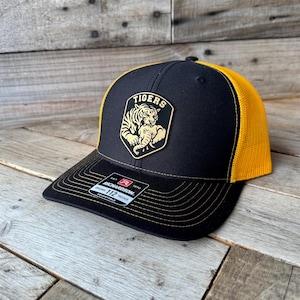 May include: Black and gold trucker hat with a tiger logo. The hat features a black front panel with a gold tiger emblem and the word "TIGERS". The mesh back is gold, and the brim is black with gold stitching.
