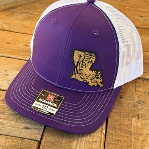 May include: Purple and white trucker hat with a gold LSU tiger logo on the front. The hat is adjustable and has a Richardson 112 label.