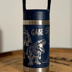 May include: A blue Yeti Rambler Jr. 14 oz. kids' water bottle with a dinosaur design and the name "GAEB" engraved on the side.