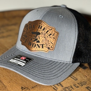 May include: A gray and black trucker hat with a brown leather patch that says "The Hell I Won't". The patch has a floral design and a cowboy silhouette.