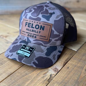 May include: A brown and black camouflage trucker hat with a brown leather patch that reads "***** FELON HILLBILLY MAKE AMERICA GREAT AGAIN! 2024". The hat has a black mesh back and a black adjustable strap with a black label that reads "EST. 1970 R RICHARDSON 112 TRUCKER ADJUSTABLE".