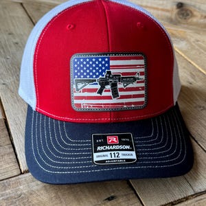 May include: Red, white, and blue trucker hat with an American flag and a rifle graphic. The hat is black on the bottom with the text "EST. 1970 RICHARDSON. ORIGINAL 112 TRUCKER ADJUSTABLE" on a white tag.