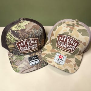 May include: Two trucker hats featuring camouflage patterns and brown mesh backs. Each hat has a rectangular patch with the text WE LIKE FAT GIRLS & FRIED BOLOGNA SANDWICHES on my momma. One hat has a dark forest camo, the other a tan camo.